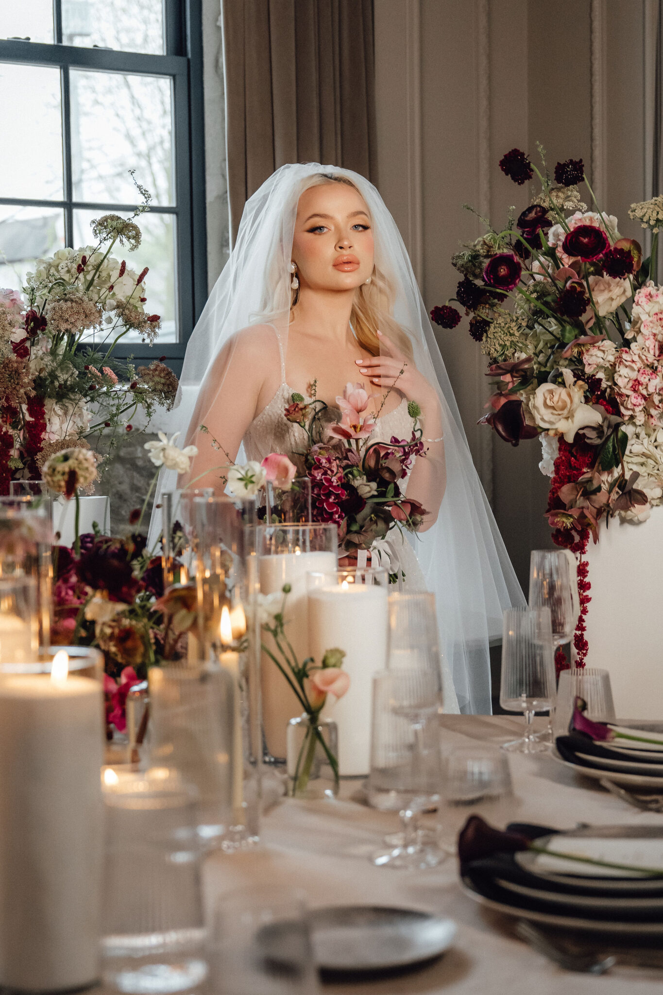 Bride in a white wedding dress and veil sits at a candlelit table surrounded by pink and burgundy flower arrangements and glassware.