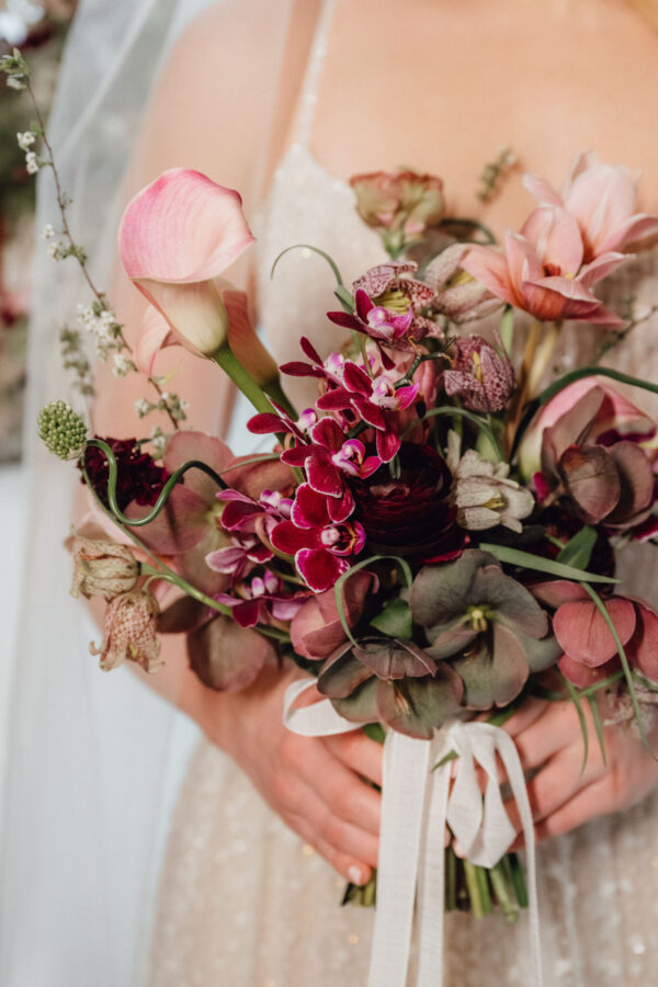 Close-up of a pink bouquet with calla lilies, orchids, and greenery held by a person in a pale dress, tied with a white ribbon