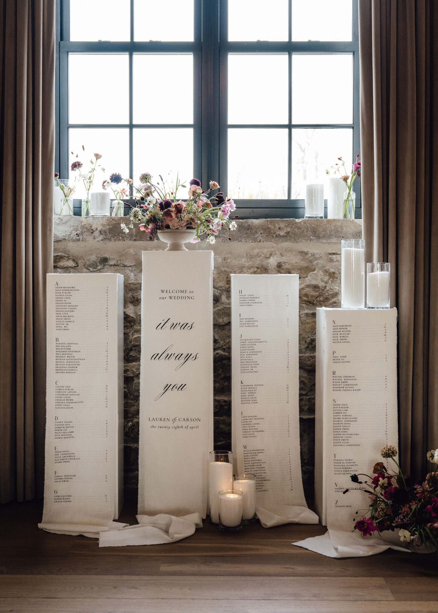 Wedding seating chart displayed on tall white panels with a floral centerpiece and candles in front of a stone wall under a window.