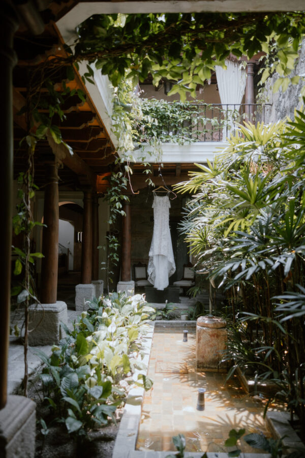 A white wedding dress hangs on a stand in a lush courtyard surrounded by green plants and vines under an overhanging balcony. Antigua, Guatemala