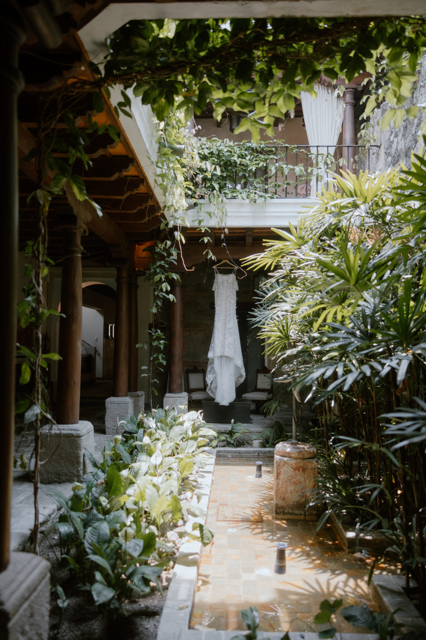 A white wedding dress hangs on a stand in a lush courtyard surrounded by green plants and vines under an overhanging balcony. Antigua, Guatemala