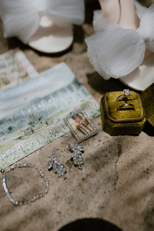 Diamond solitaire engagement ring in a green velvet box, surrounded by jewelry on a sandy surface.