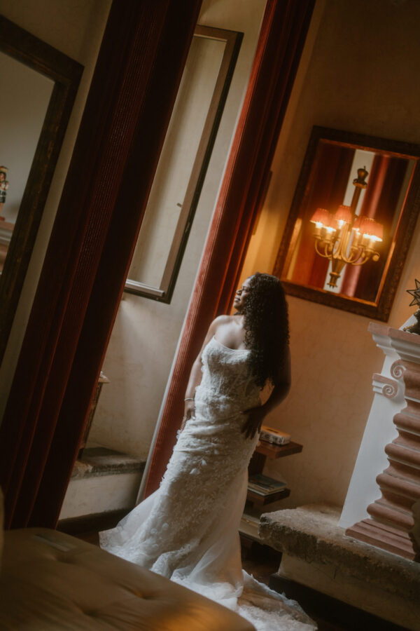 Brid​e in a white lace wedding gown standing in a warmly lit, vintage-style room with wooden mirrors and a chandelier.
