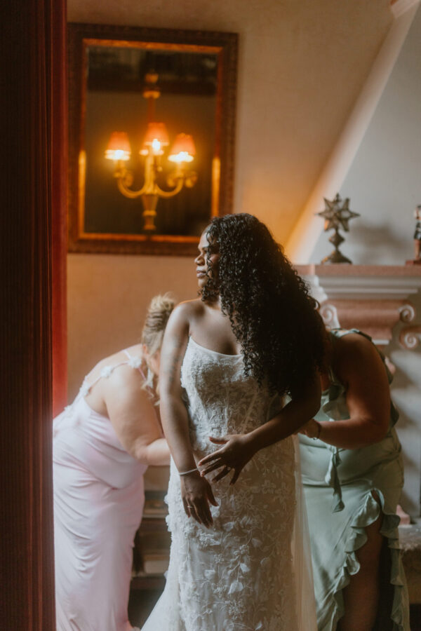 Bride in a lace wedding gown stands in a warm, chandelier-lit room as bridesmaids help her dress