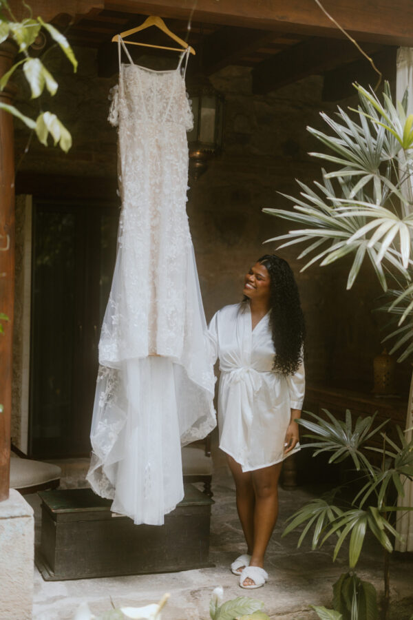 Wedding dress on a hanger as a woman in a satin robe smiles at it in a plant-filled courtyard.