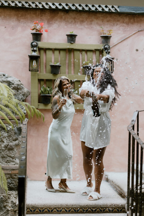 Two women in white dresses toast with champagne as bubbles spray through the air on a decorated stairway.