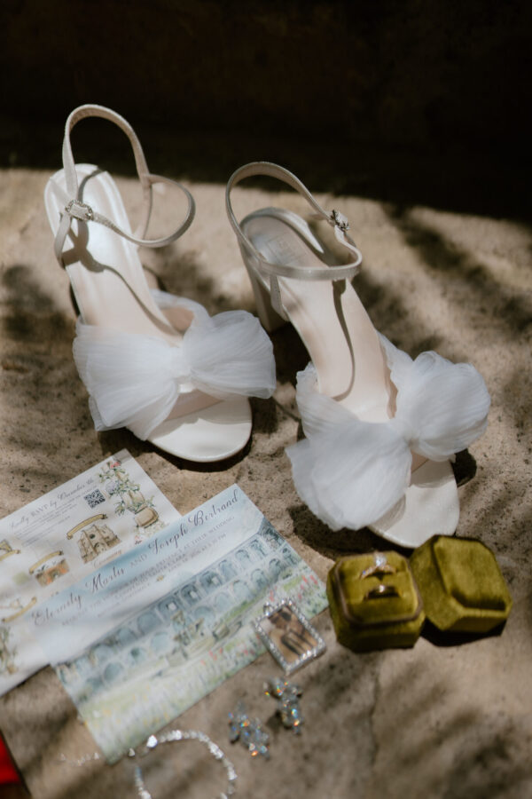 White bridal heels with delicate straps and tulle embellishments resting on sandy ground with wedding invitations nearby.