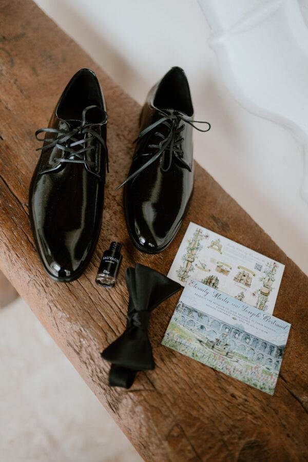 Pair of shiny black dress shoes with laces on a wooden surface beside a bow tie and illustrated wedding invitation cards.