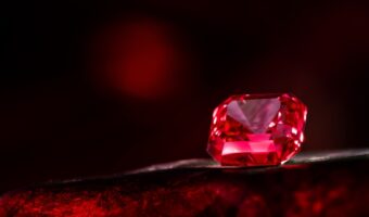 Close-up of a faceted red gemstone resting on a dark red surface.
