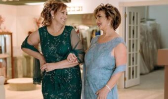 Two women in formal dresses smile at each other while standing arm-in-arm in a boutique, one in green and the other in light blue.