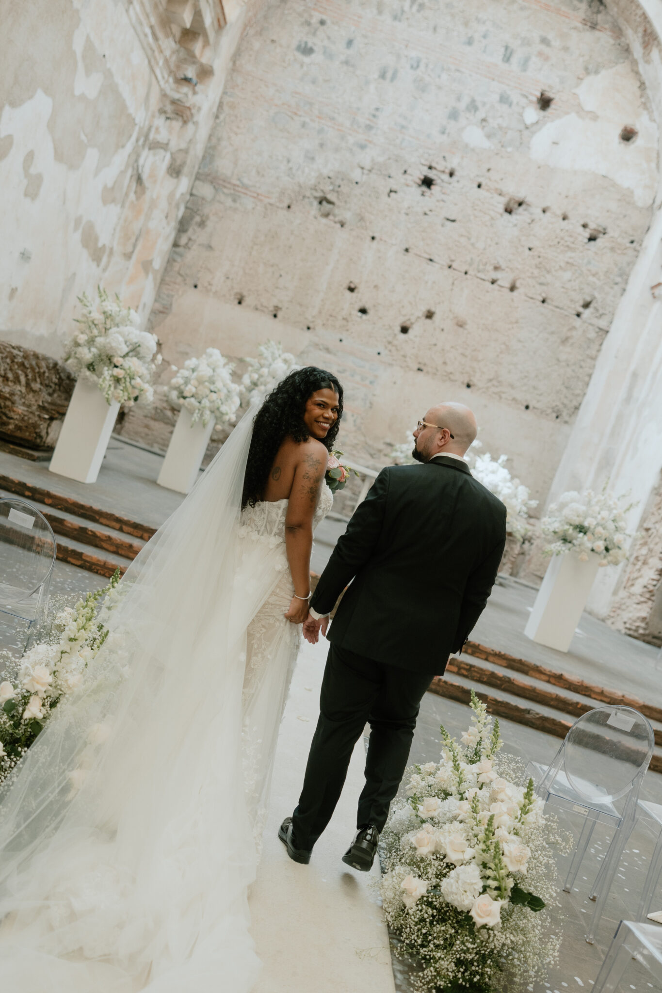 Bride in lace wedding gown with long veil smiling at groom as they walk down a floral‑decorated aisle in a rustic venue with exposed brick walls on either side.Antigua, Guatemala