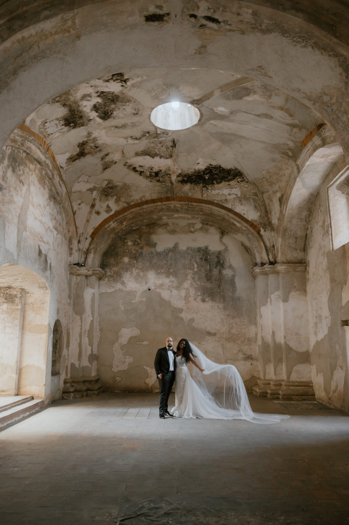 Bride and groom stand together in a weathered stone hall with soaring arches and a circular skylight above them, wedding veil trailing. Antigua, Guatemala