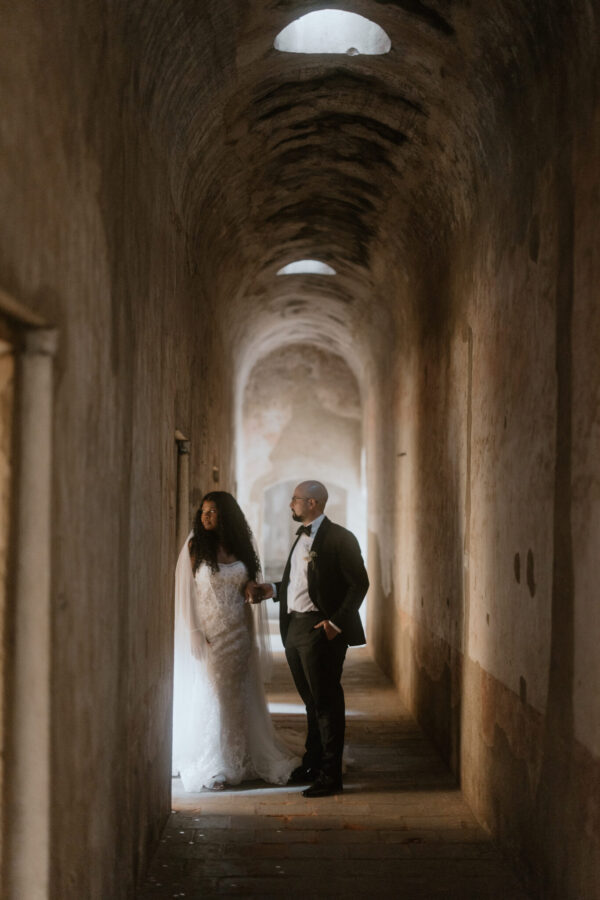 Bride in a white wedding gown and groom in a suit stand hand in hand in a dim, arched stone corridor.