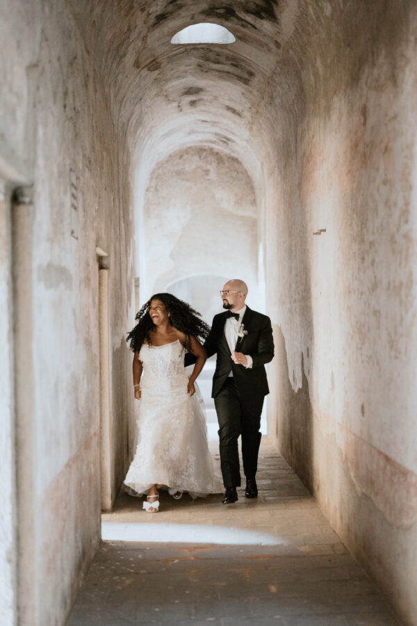 A bride in a lace wedding dress and a groom in a black suit walk arm in arm down a dusty, arched stone hallway.