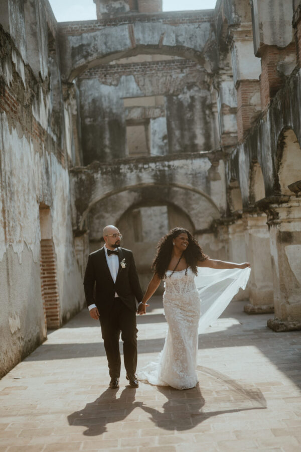 Bride in a lace wedding gown and groom in a black tuxedo walk hand in hand through ancient stone arches in a ruins setting.