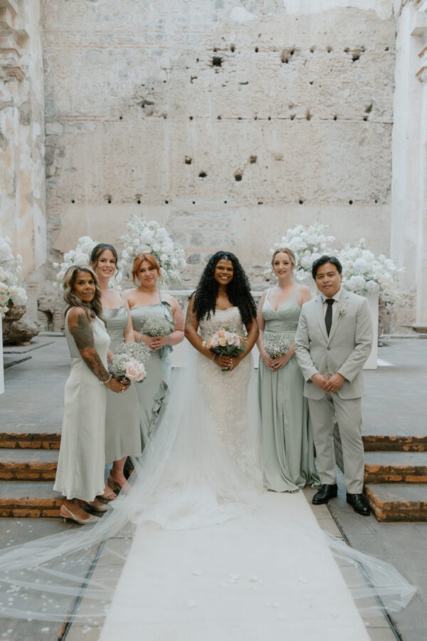Bride in a white lace gown stands at the center with a bouquet, flanked by bridesmaids in sage dresses and a man in a light gray suit, on a stone stairway with white floral arrangements behind them.