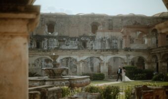 Couple in wedding attire walking through a historic stone courtyard with arches and a central fountain at Antigua, Guatemala