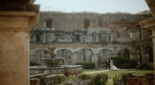 Couple in wedding attire walking through a historic stone courtyard with arches and a central fountain at Antigua, Guatemala