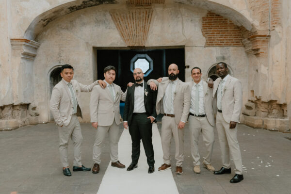 Group of seven men in light gray suits posing together outdoors in front of a rustic stone archway. One man wears a black suit and bow tie.