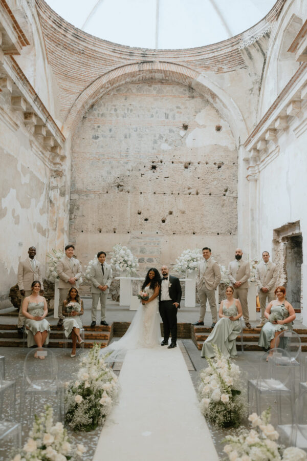 Bride and groom stand at a white aisle altar inside a ruined stone chapel, surrounded by bridesmaids and groomsmen in pale beige attire.