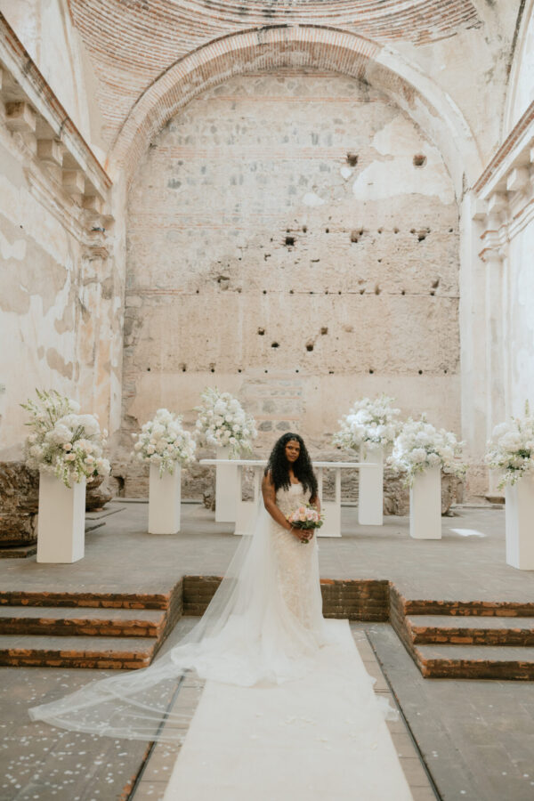 Bride in a lace wedding dress holding a bouquet, standing on a white aisle with a long veil in a historic stone venue.