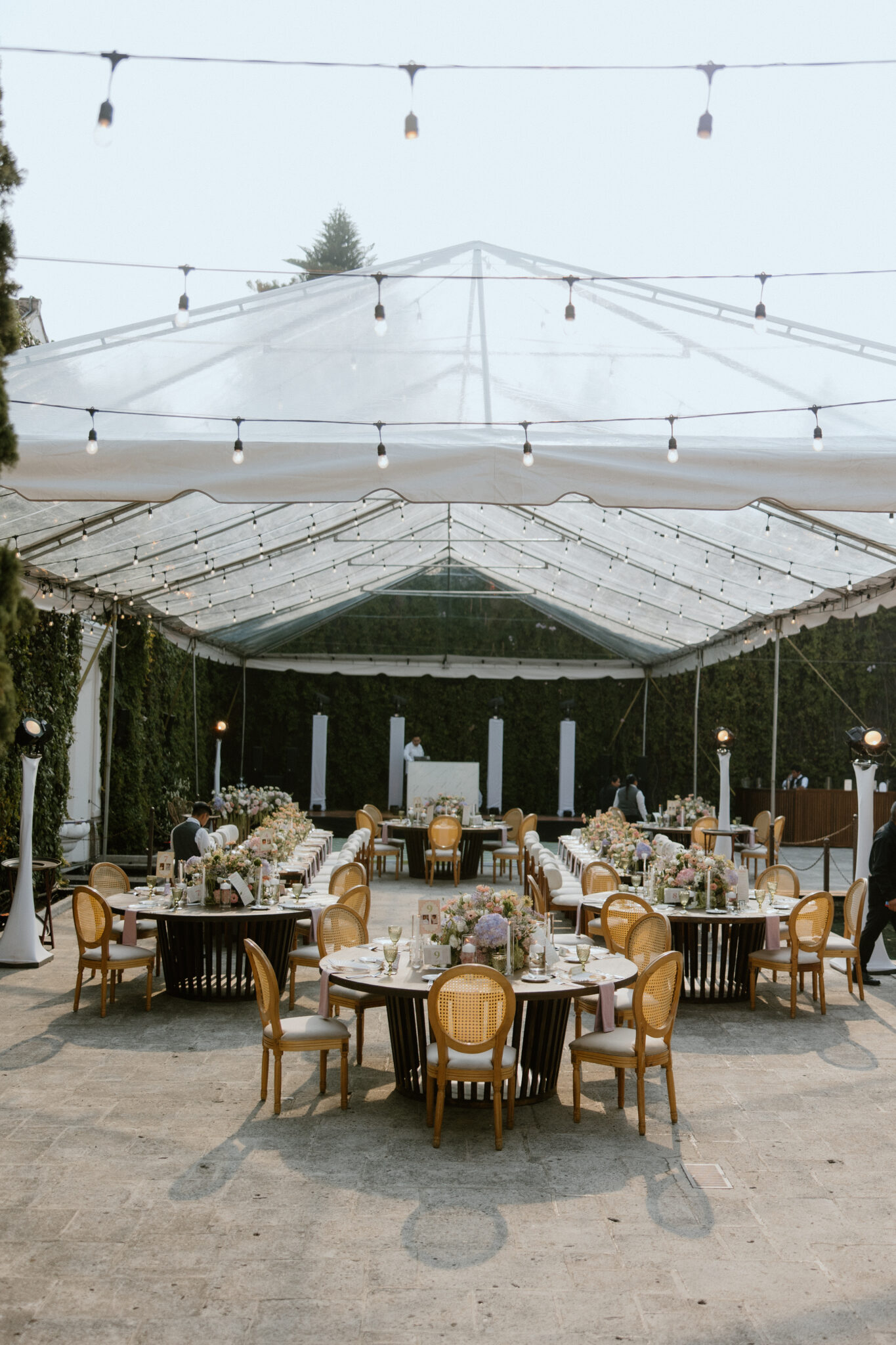 Outdoor wedding reception under a clear tent with string lights, long banquet tables adorned with pink and white floral centerpieces and cane-back chairs.