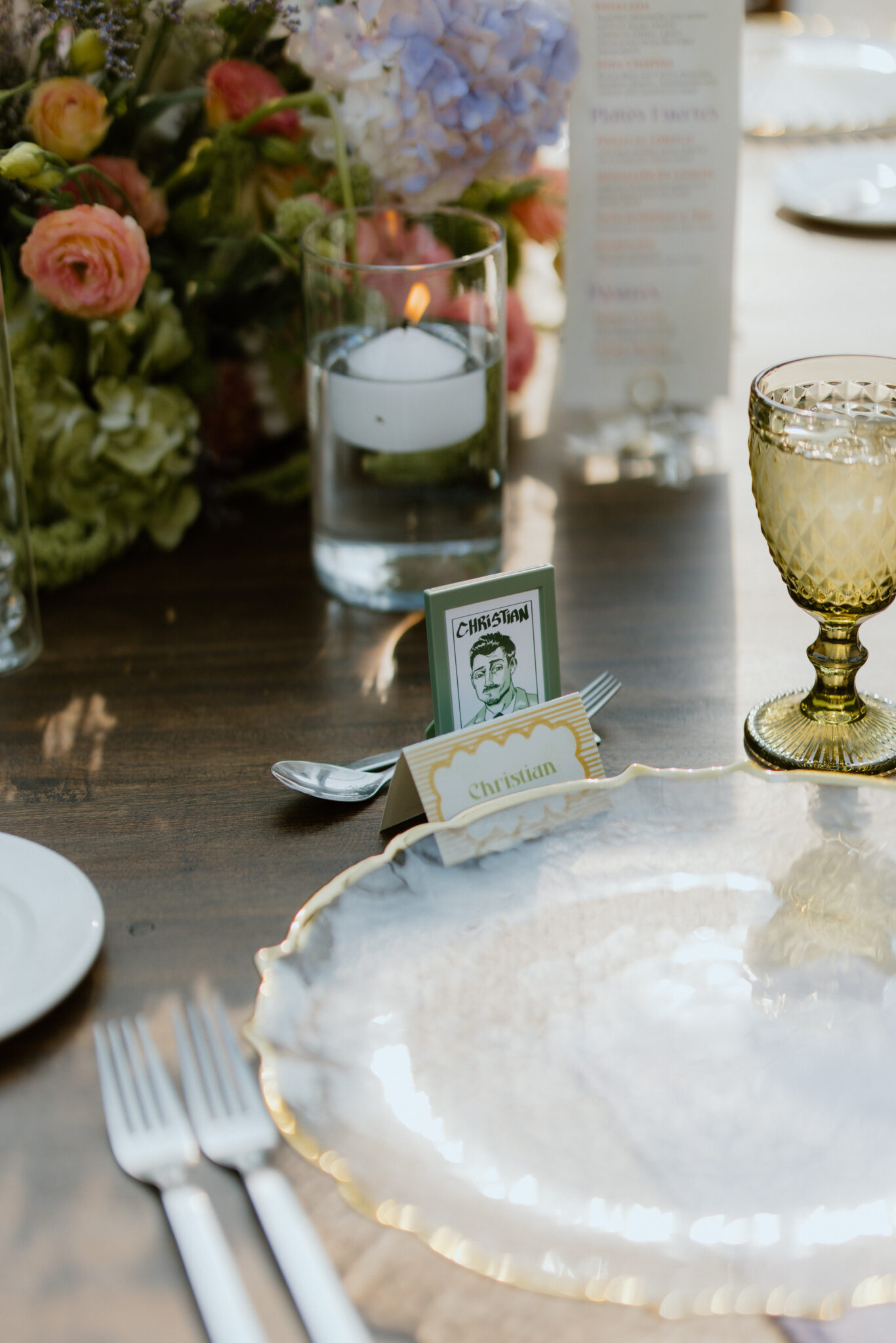 Close-up of an elegant table setting with a lit candle, pastel flowers, a gold-rimmed plate, and a place card reading Christian.