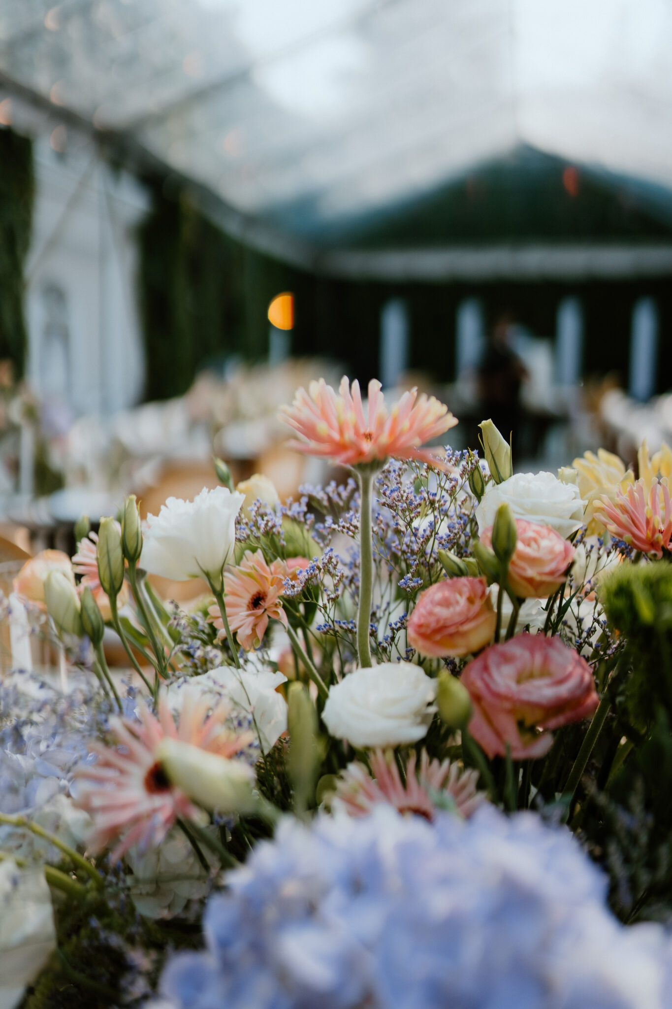 Close-up of a pastel flower bouquet—pink, peach, white blooms with blue hydrangeas—at a banquet table, background blurred into soft tones.