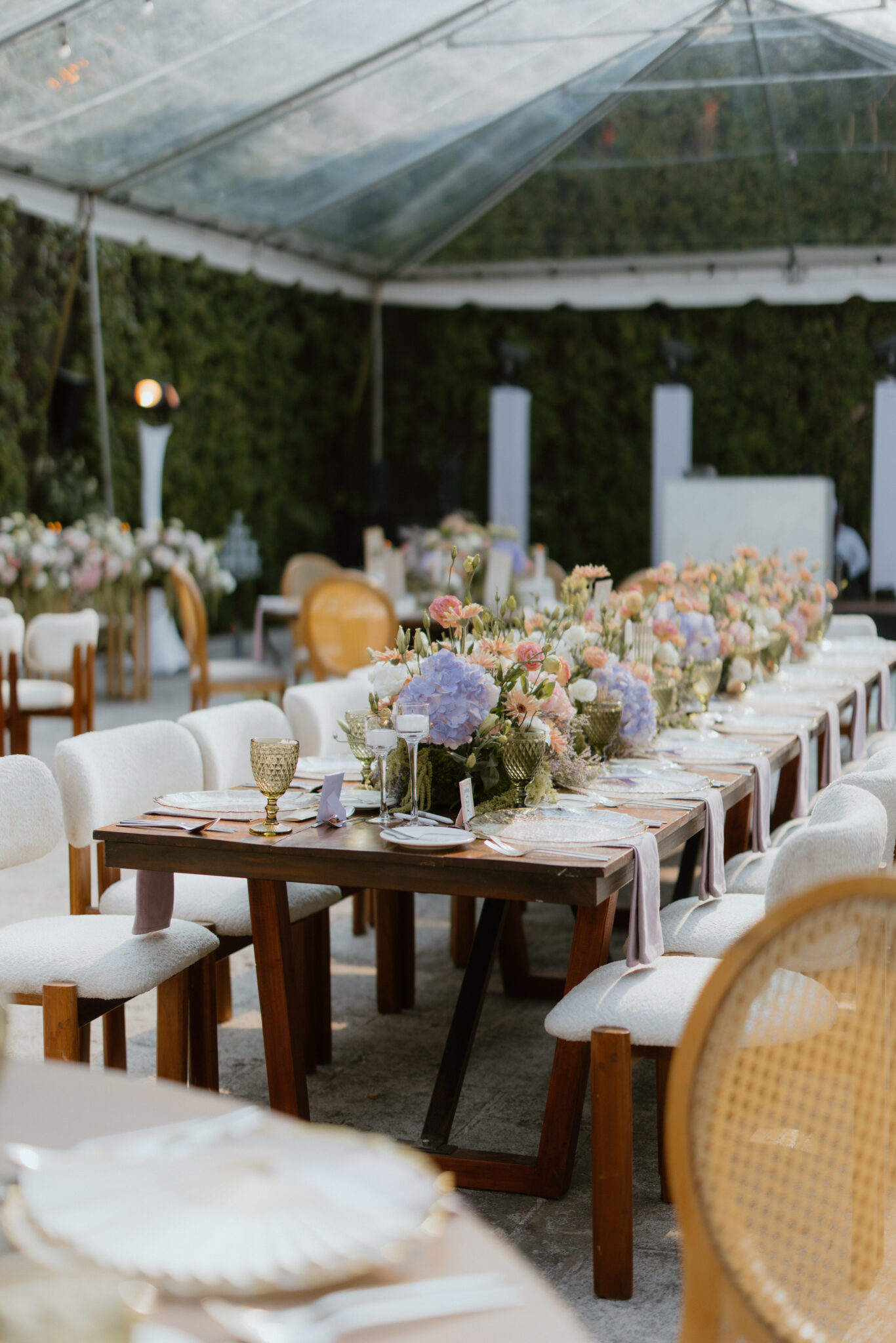 Long wooden banquet table with pastel floral centerpieces under a glass tent, set with plates, glasses, and pink napkins alike for a wedding reception.