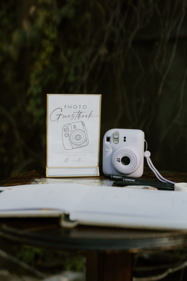 White instant camera beside a 'Photo Guestbook' sign on a wooden table, with pens and papers nearby outdoors.