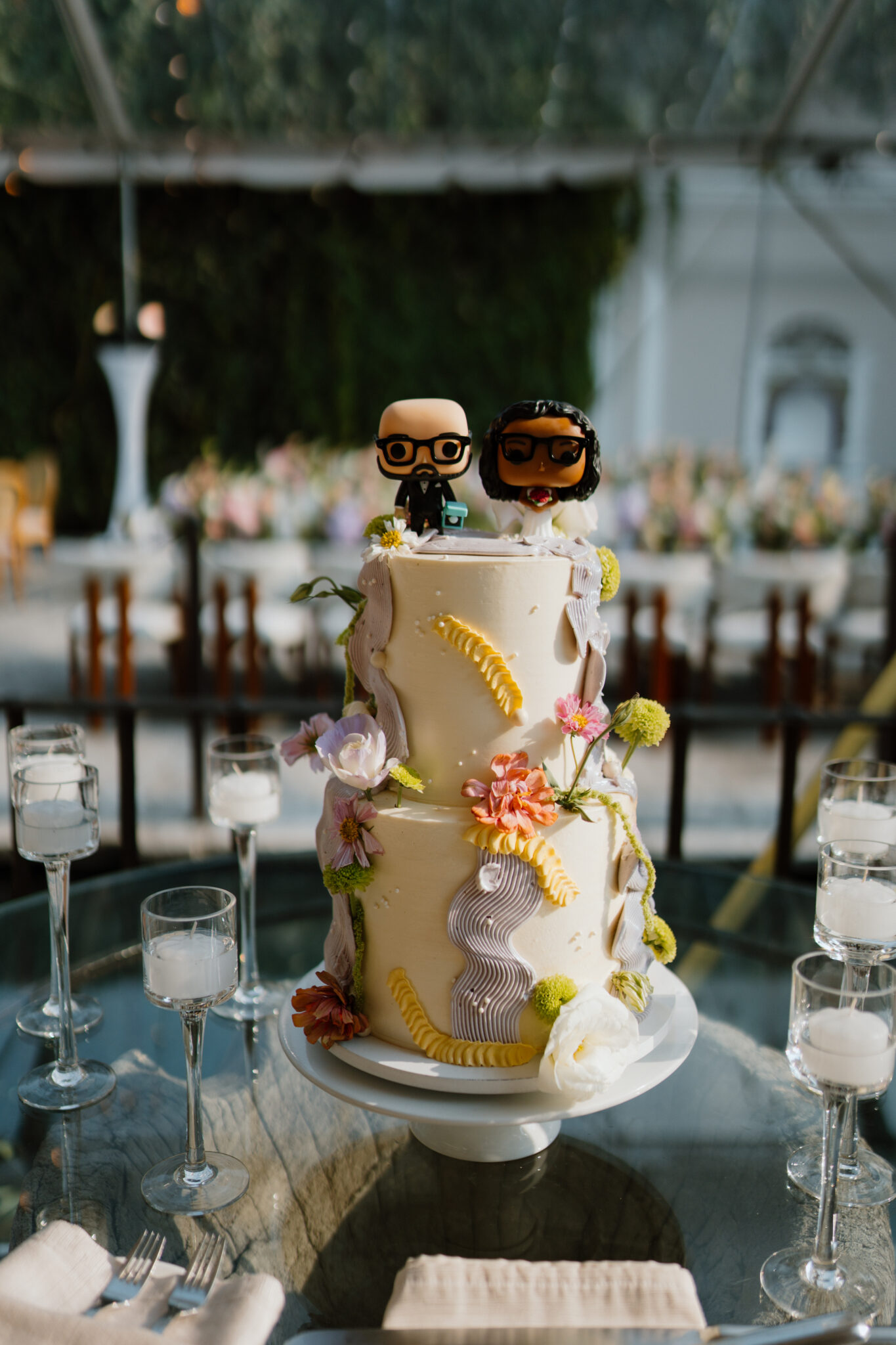 Wedding cake with two figurine toppers wearing glasses on top, surrounded by pastel flowers on a glass table with candles in the foreground.