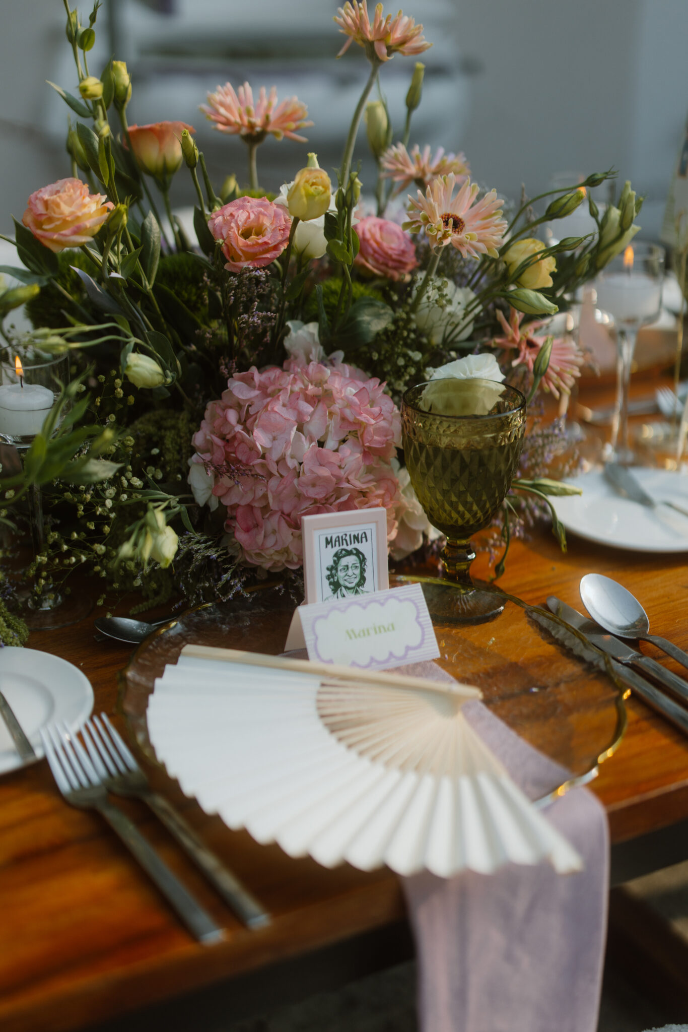 Floral centerpiece with pink and peach flowers on a wooden table, surrounded by place settings and candles.