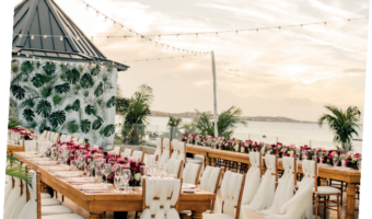 Long wooden wedding table on a waterfront terrace, with white chairs, pink flowers, palm plants, and string lights at sunset.