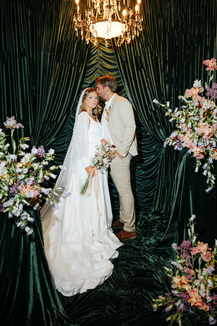 Bride in a white wedding gown and veil stands beside a groom in a beige suit under a chandelier, framed by dark green velvet drapes and floral arrangements.
