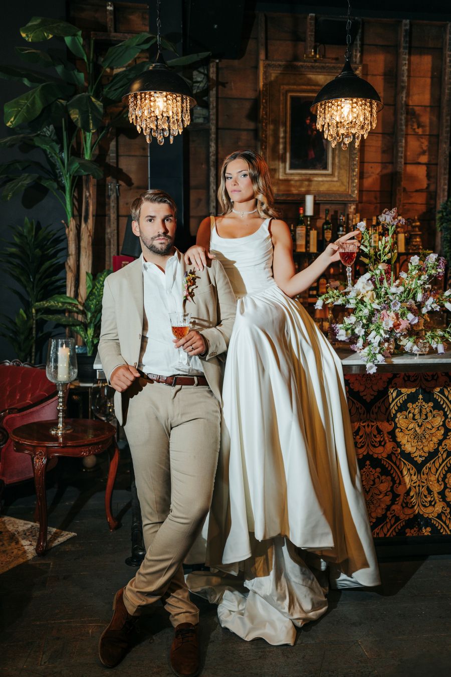 Man in a beige suit and a woman in a white gown pose at a stylish bar, each holding a cocktail.