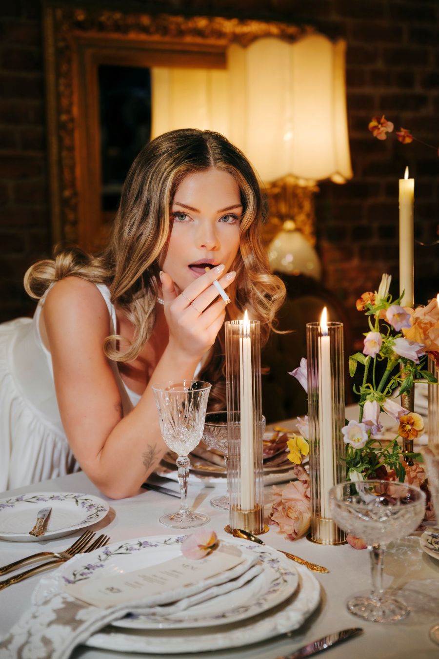A woman in a white dress smokes a cigarette at a candlelit, flower-adorned dining table.