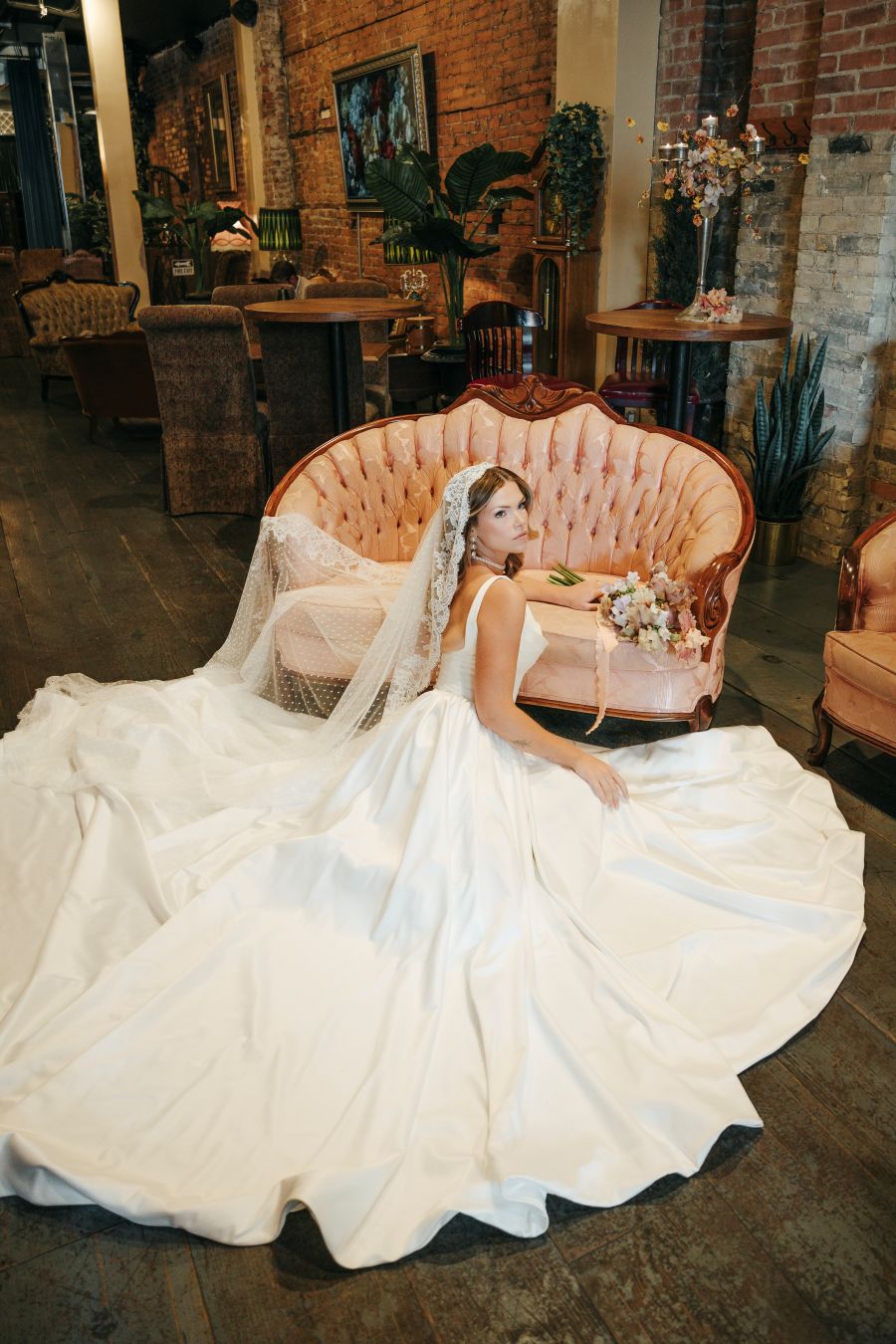 Bride in a white wedding gown and veil sits on a pink tufted sofa, holding a bouquet in a vintage-inspired indoor venue with exposed brick walls.