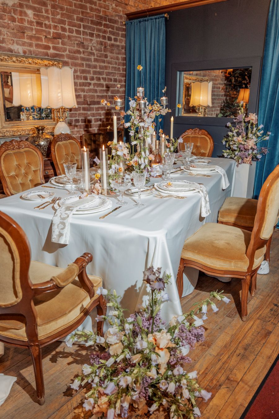Elegant dining table set for a formal dinner with candles, floral centerpieces, crystal glassware, and vintage chairs in a brick-walled room and blue drapes.