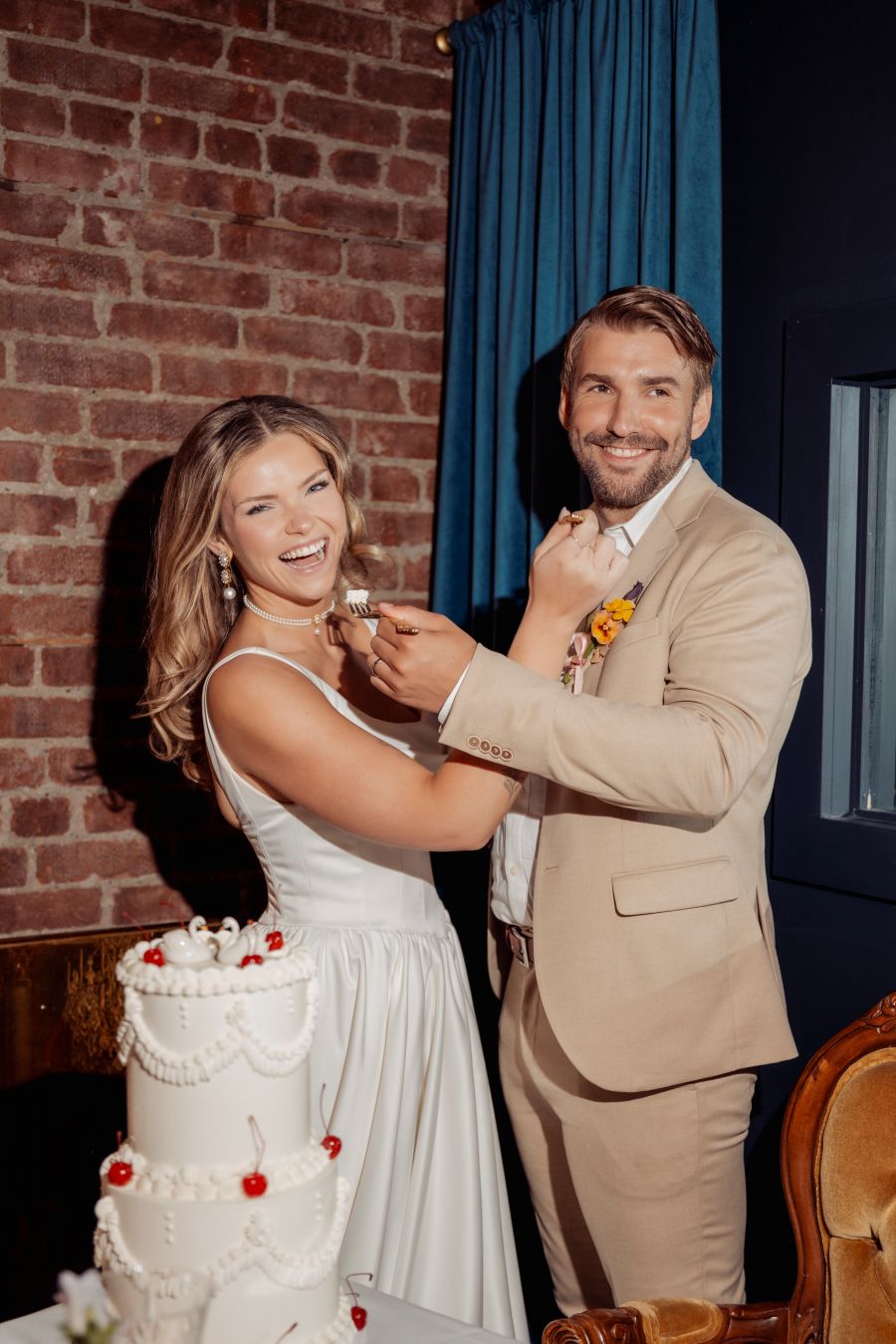Bride and groom cutting and feeding each other wedding cake in a rustic brick venue.