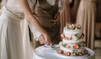 Bride and partner slice a three-tier wedding cake decorated with strawberries and roses on a white-tablecloth table.