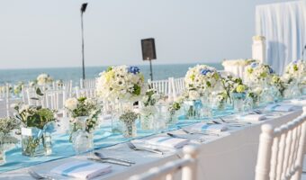 Long seaside wedding table with white chairs, blue runners, and glass flower centerpieces overlooking the ocean at sunset/sunlit day.