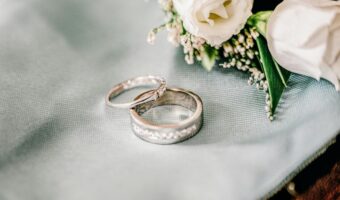 Two wedding bands, one plain silver and one diamond-studded, lie on pale blue fabric with a white bouquet nearby.
