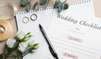 Wedding checklist on a desk with a pen, rings, ribbon, and white flowers.