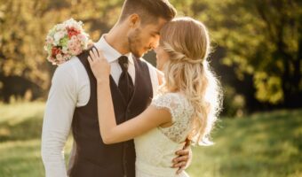 Bride and groom in wedding attire embrace in a sunlit park, the bouquet resting on the groom's shoulder as they lean toward each other.