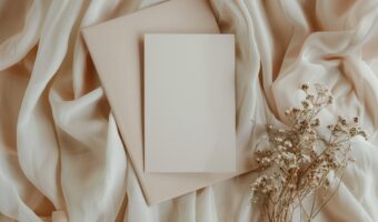 Flat lay of a blank white card on a beige envelope, surrounded by cream fabric and dried flowers.