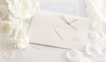 Elegant white wedding card with an embossed heart and bow, surrounded by white roses and petals.
