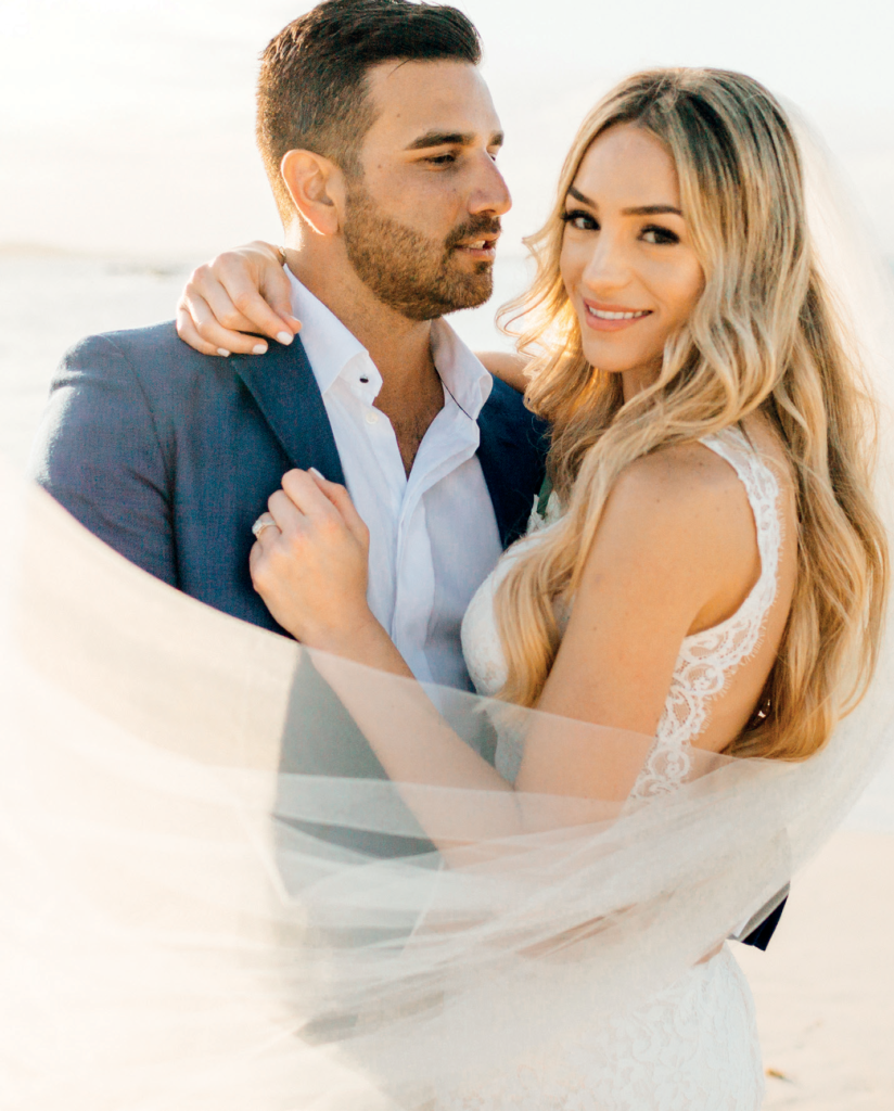 Couple on a sunny beach: bride in a lace wedding dress and veil embraces a man in a navy suit, smiling at the camera with warm light