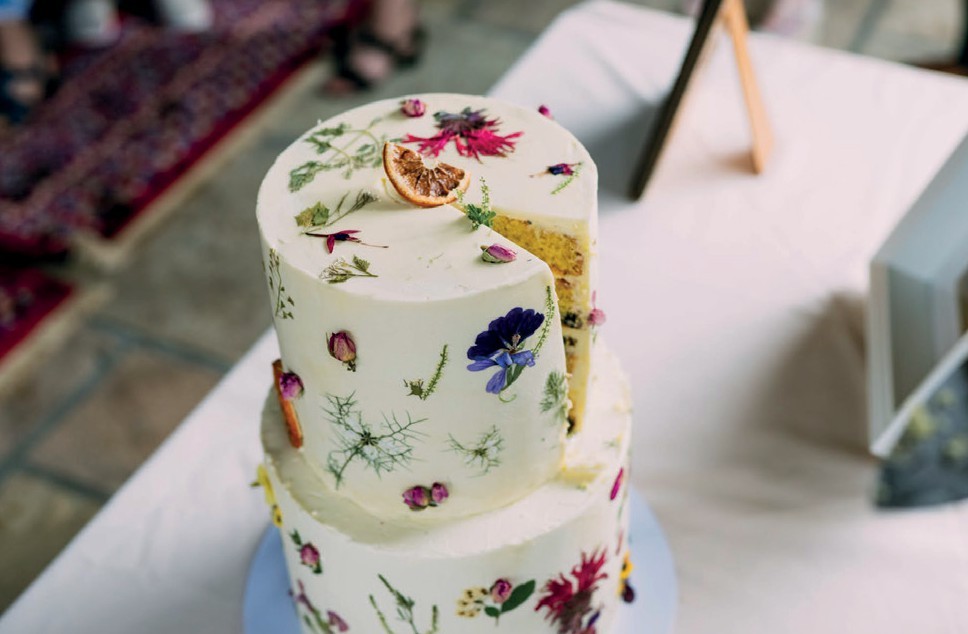 Two-tier white cake decorated with pressed flowers; a slice is removed to reveal the yellow interior on a light table.