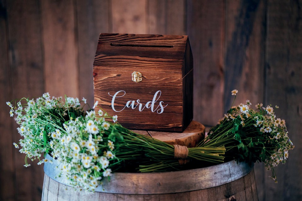 Wooden card box labeled 'Cards' sits on a rustic barrel, framed by white daisies and greenery on a wooden backdrop.