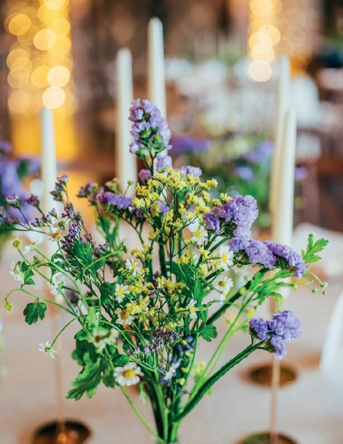 Centerpiece of colorful wildflowers in a glass vase on a dining table, with tall cream candles and soft warm bokeh lights in the background